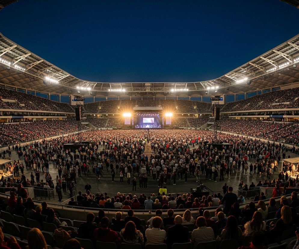 Ogromny stadion jest wypełniony tłumem ludzi, skupionych na scenie umieszczonej na jednym z końców płyty boiska. Scena oświetlona jest jasnym, ciepłym światłem, kontrastującym z ciemnogranatowym niebem nad otwartym dachem obiektu. Widownia rozmieszczona jest zarówno na płycie, jak i na trybunach, tworząc falę ludzkich sylwetek, z których część na pierwszym planie jest dobrze widoczna, a dalsze sekcje zlewają się w masę. Górna część stadionu, wraz z dachem i elementami konstrukcyjnymi, jest również podświetlona, co dodaje głębi scenie.