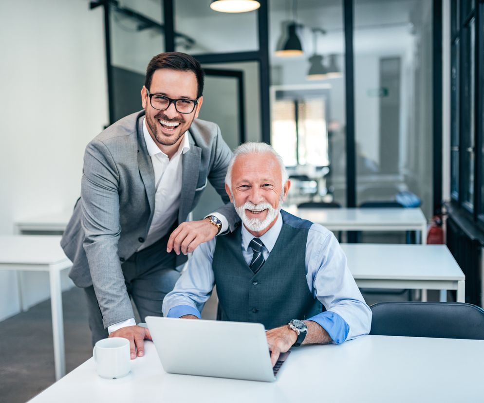 Uśmiechnięci mężczyźni w biurze: młodszy, w szarym garniturze, opiera się o ramię starszego, eleganckiego mężczyzny z siwą brodą, siedzącego przy laptopie. Obaj zadowoleni, symbolizujący pracę po emeryturze i korzyści z jej odkładania, o czym możesz przeczytać na Super Biznes.