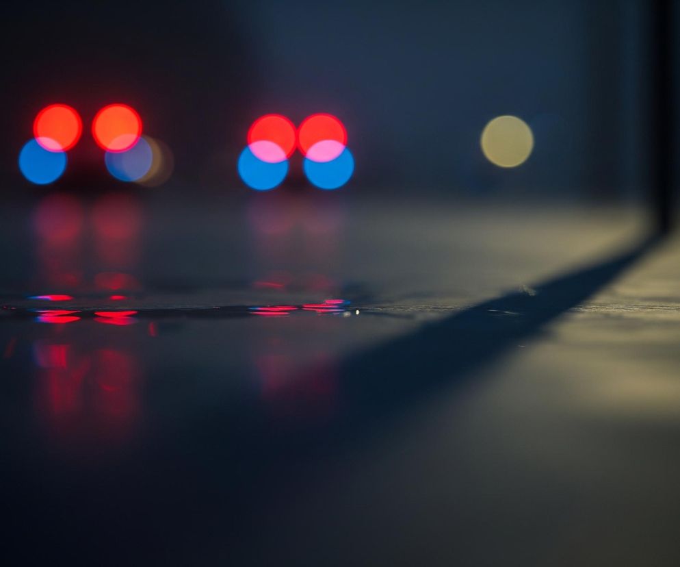 Rozmyte, kolorowe światła rozświetlają ciemne tło. Na pierwszym planie widoczne są odbicia tych świateł w niewielkich kałużach wody na ciemnej powierzchni. Po lewej stronie znajdują się dwa duże czerwone i dwa mniejsze niebieskie światła, z których każde ma swoje odbicie w dole. Po prawej stronie w oddali widoczne jest pojedyncze, żółtawe, rozmyte światło. Ciemny cień przecina ukośnie powierzchnię od prawej strony kadru ku górze.