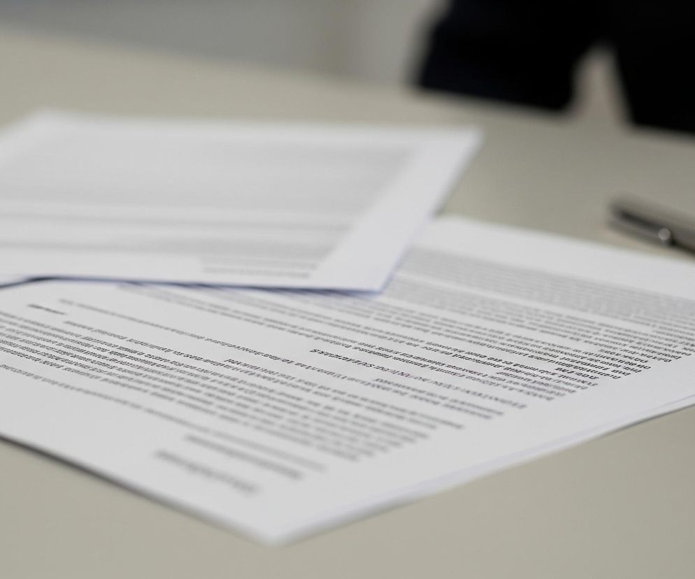 Na jasnoszarym blacie leżą dwie kartki papieru. Jedna, ostra i wyraźna, zajmuje większą część kadru, przedstawiając gęsty tekst pisany czarną czcionką, z nagłówkiem Dienstübergabe w lewym górnym rogu. Druga kartka, rozmyta, znajduje się za nią, częściowo zasłonięta, również pokryta tekstem. Na prawej stronie ostrej kartki, w pobliżu górnej krawędzi, leży srebrny długopis z czarnym elementem, częściowo wyostrzony. Tło jest rozmyte, ukazując niewyraźny kształt w ciemnym kolorze, prawdopodobnie fragment ubrania lub innego obiektu.