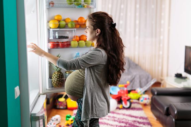 Kobieta w zaawansowanej ciąży stojąca przed otwartą lodówką