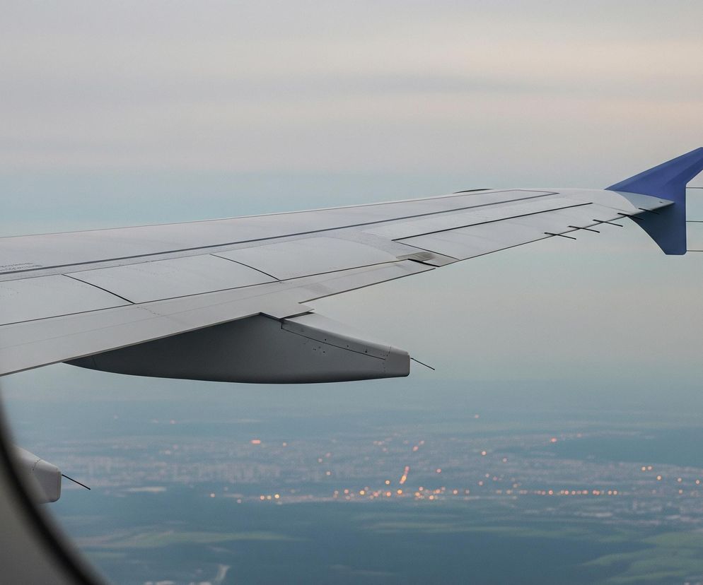 Lewa strona kadru jest przysłonięta ciemną, pionową krawędzią okna samolotu. Przez okno widać skrzydło samolotu w odcieniach szarości, rozciągające się ukośnie od lewego dolnego rogu ku prawej górze, z charakterystycznym niebieskim zakończeniem (wingletem). Poniżej skrzydła rozciąga się rozmyty krajobraz, częściowo pokryty zielonymi i szarymi obszarami, z licznymi drobnymi, żółto-pomarańczowymi punktami świetlnymi, przypominającymi światła miasta po zmierzchu. Górna część nieba jest jasna, w odcieniach szarości i błękitu, przechodząc w jaśniejszy horyzont.