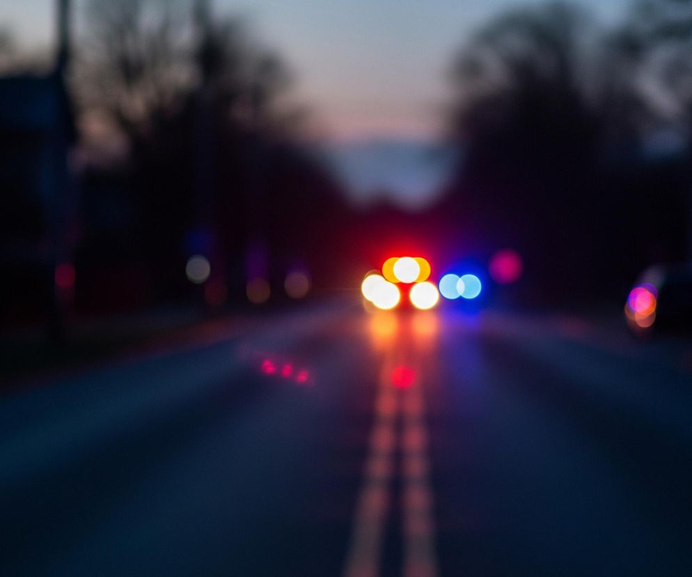 Na środku rozmytego zdjęcia widać pojazd z włączonymi, rozmazanymi światłami w kolorach czerwonym, pomarańczowym i niebieskim. Światła te rzucają poświatę na ciemną nawierzchnię drogi, która jest przedzielona dwoma równoległymi, jasnymi liniami. Po obu stronach drogi, w oddali, znajdują się niewyraźne, ciemne kształty, przypominające drzewa lub budynki. Niebo w tle jest w odcieniach szarości i błękitu, z delikatnymi różowymi akcentami przy horyzoncie.