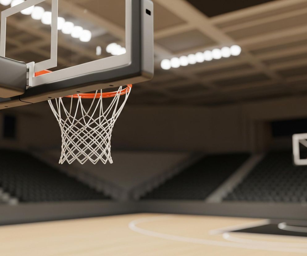 Na pierwszym planie, z lewej strony, widoczna jest obręcz do koszykówki z białą siatką, przymocowana do ciemnoszarej tablicy. Pod obręczą znajduje się jasnobrązowy parkiet boiska z białymi liniami. W tle, na wprost, widać trybuny z rzędami czarnych siedzeń, a za nimi kolejną, oddaloną obręcz do koszykówki. Sufit hali sportowej jest beżowy, z widocznymi, równomiernie rozmieszczonymi jasnymi punktami świateł.