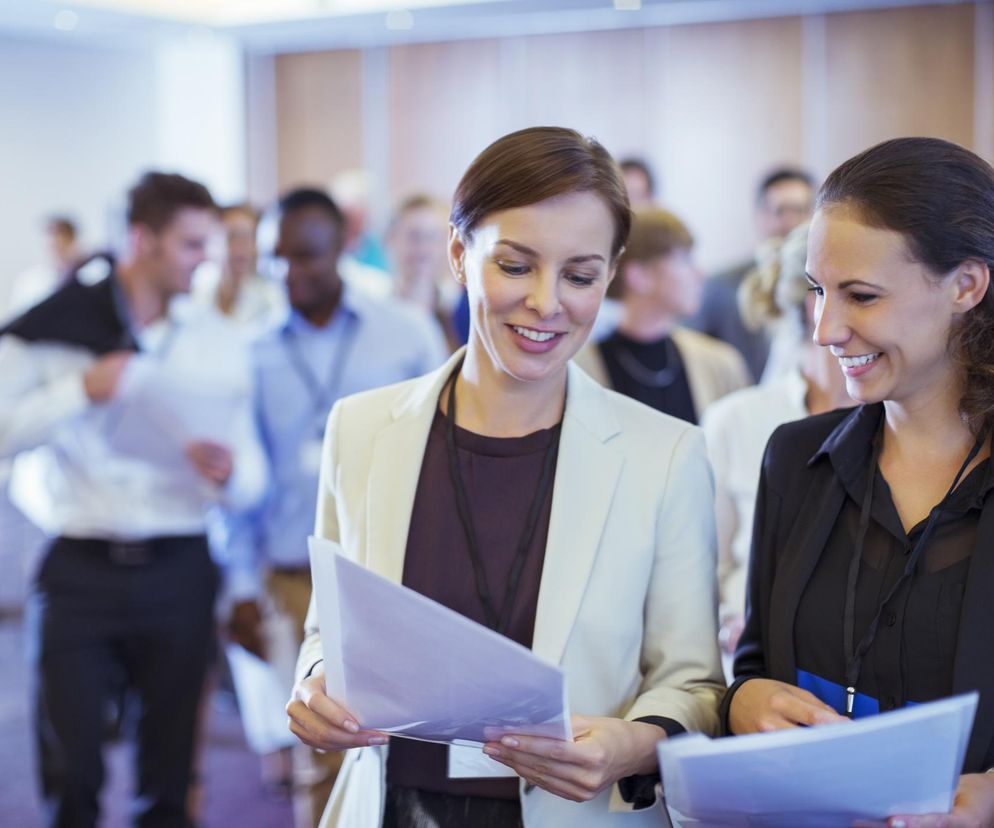 Dwie kobiety w strojach biznesowych uśmiechają się, trzymając w rękach dokumenty. Stoją w tłumie, co odzwierciedla kontekst artykułu o ryzyku rozprzestrzeniania się infekcji podczas głośnych rozmów w zatłoczonych miejscach. O tym, jak chronić się przed zarazkami, przeczytasz na Poradnik Zdrowie.