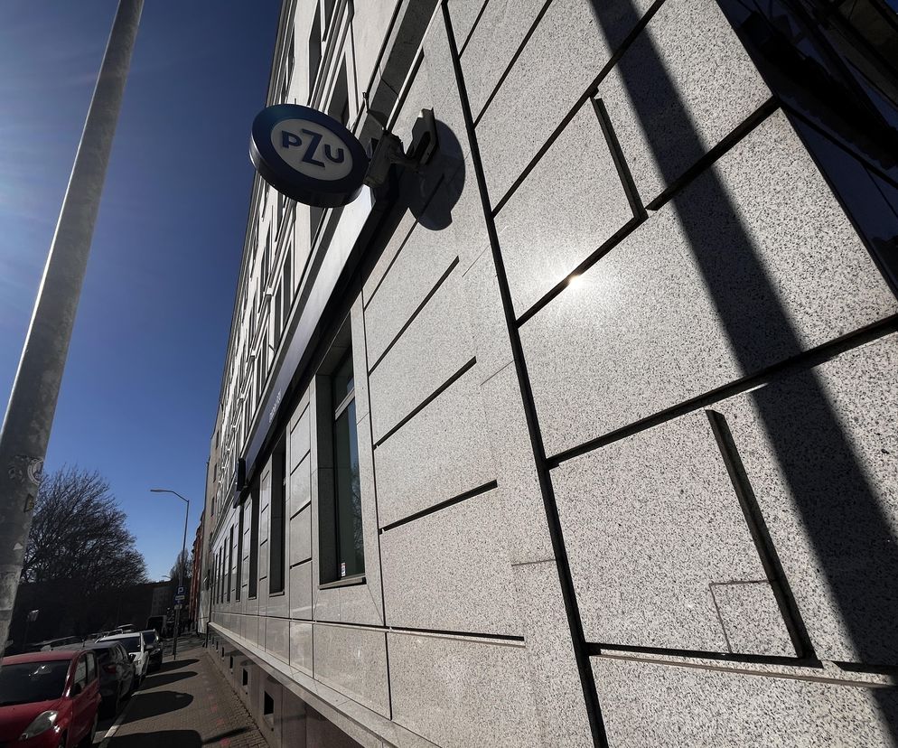 Znak z logo PZU na fasadzie budynku w Szczecinie. Słońce odbija się w ścianach i rzuca długie cienie na elewację, co wizualnie oddaje kontekst artykułu o finansowych roszczeniach Skarbu Państwa wobec byłej prezes PZU. Więcej na Super Biznes.