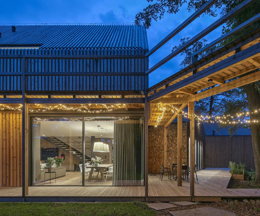 Nowoczesny budynek mieszkalny Polska Zagroda BXB Studio w Bulowicach z drewnianą elewacją i przeszkleniami. Wieczorna sceneria z oświetleniem tarasów, podkreślająca integrację architektury z otoczeniem. Więcej o tym projekcie na Architektura Murator Plus.