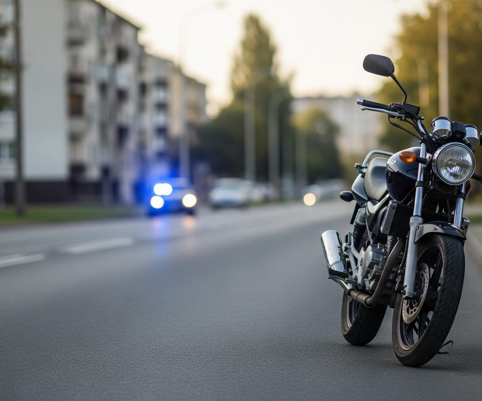Czarny motocykl, stojący na poboczu drogi, z perspektywą lekko od przodu, dominuje prawą stronę obrazu. Jego przednie koło znajduje się na asfalcie, a reszta pojazdu stoi na tle ulicy i rozmytemu tłu miejskiego krajobrazu. Na tle rozmytych drzew i budynków, w oddali widoczne są niebieskie światła pojazdu, prawdopodobnie radiowozu policyjnego, oraz inne, niewyraźne światła samochodów. Ulica, z widocznymi białymi liniami, rozciąga się w lewą stronę zdjęcia, podkreślając głębię obrazu.