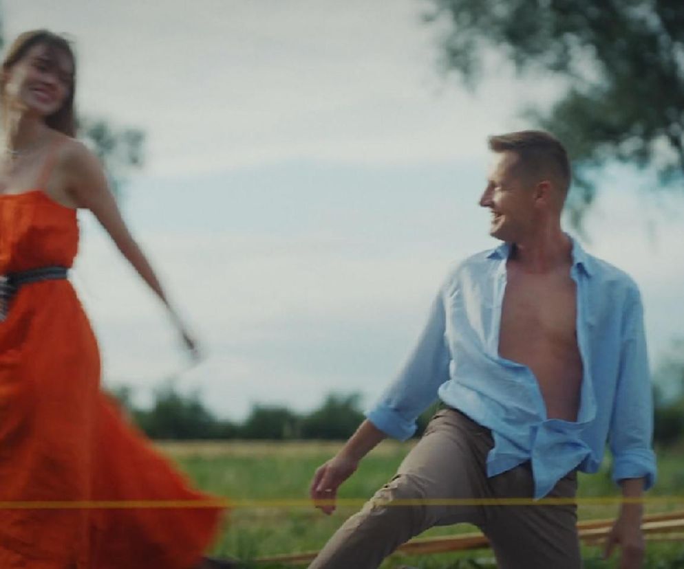 Dominika Kachlik (Franka) w zwiewnej, pomarańczowej sukience i Rafał Mroczek (Paweł) z rozpiętą koszulą na tle zieleni, uśmiechający się do siebie, symbolizujący romantyczny piknik i rodzącą się namiętność w jubileuszowym odcinku M jak miłość. Więcej o ich losach na Super Seriale.