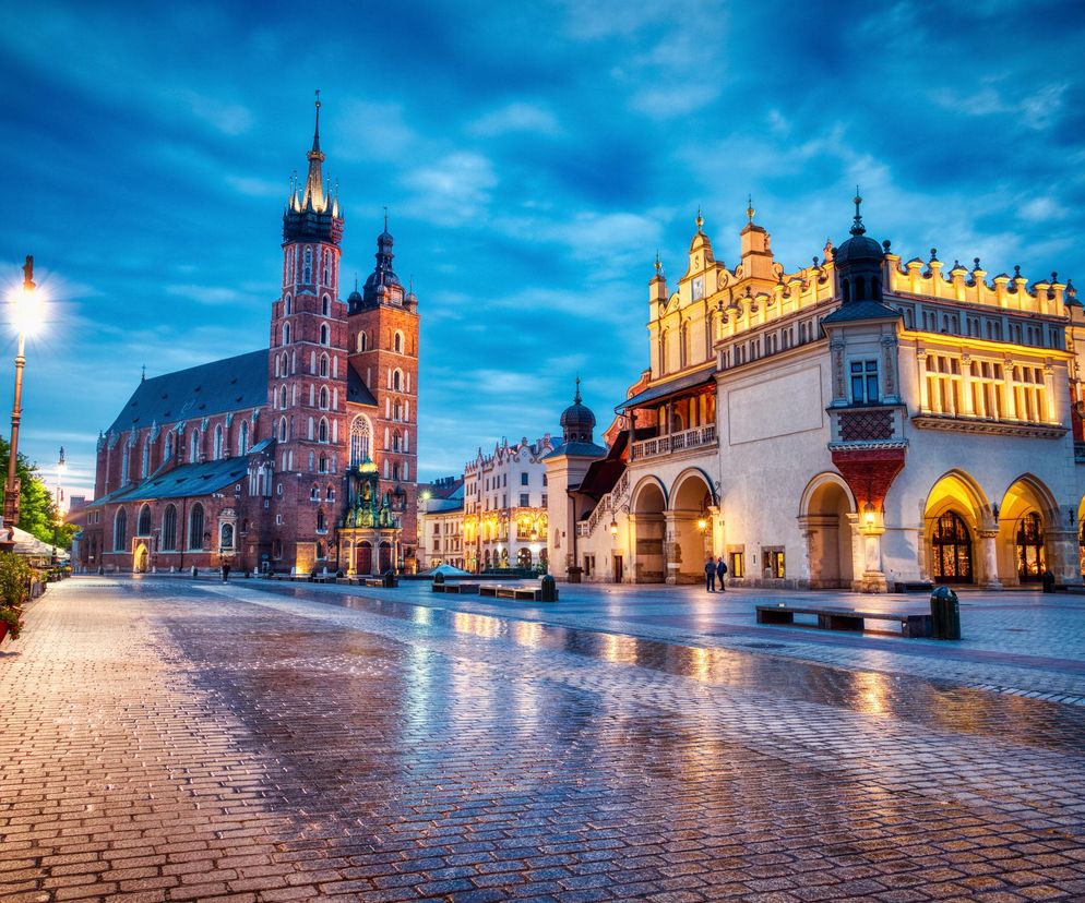 Rynek Główny w Krakowie o zmierzchu, z widocznym Kościołem Mariackim po lewej i Sukiennicami po prawej stronie. Brukowany plac odbija światła latarni i oświetlonych budynków. To zdjęcie ilustruje ranking miast, gdzie żyje się najlepiej, o którym możesz przeczytać na Poradnik Zdrowie.