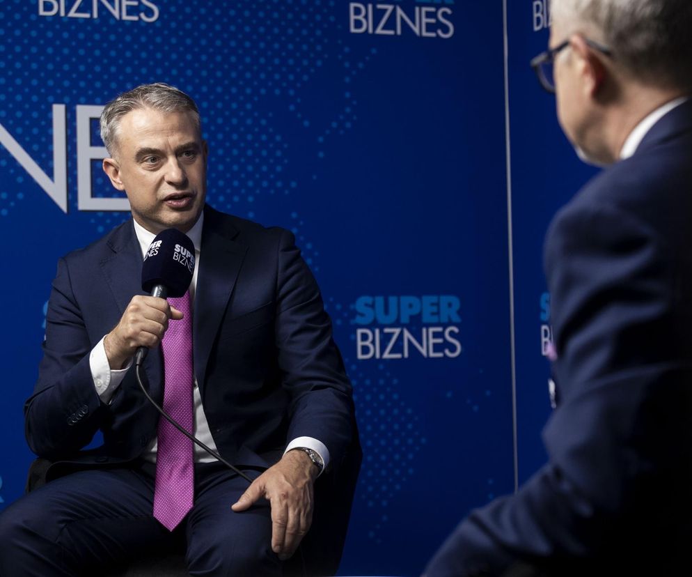 Wicepremier Krzysztof Gawkowski podczas wywiadu na tle logo Super Biznes, trzymający mikrofon z logo Super Biznes, rozmawiający z niewidoczną osobą. Na Super Biznes można przeczytać o cyberzagrożeniach i rekomendacjach ministra.