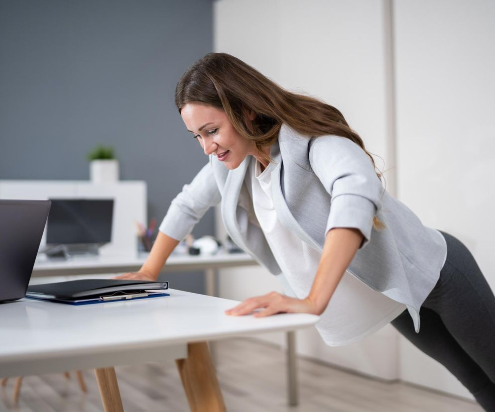 Kobieta w białej bluzce i szarym żakiecie wykonuje ruchową przekąskę – pompkę na biurku, co ma pozytywny wpływ na koncentrację i zdrowie serca. Więcej o prostych ćwiczeniach przeczytasz na Poradnik Zdrowie.