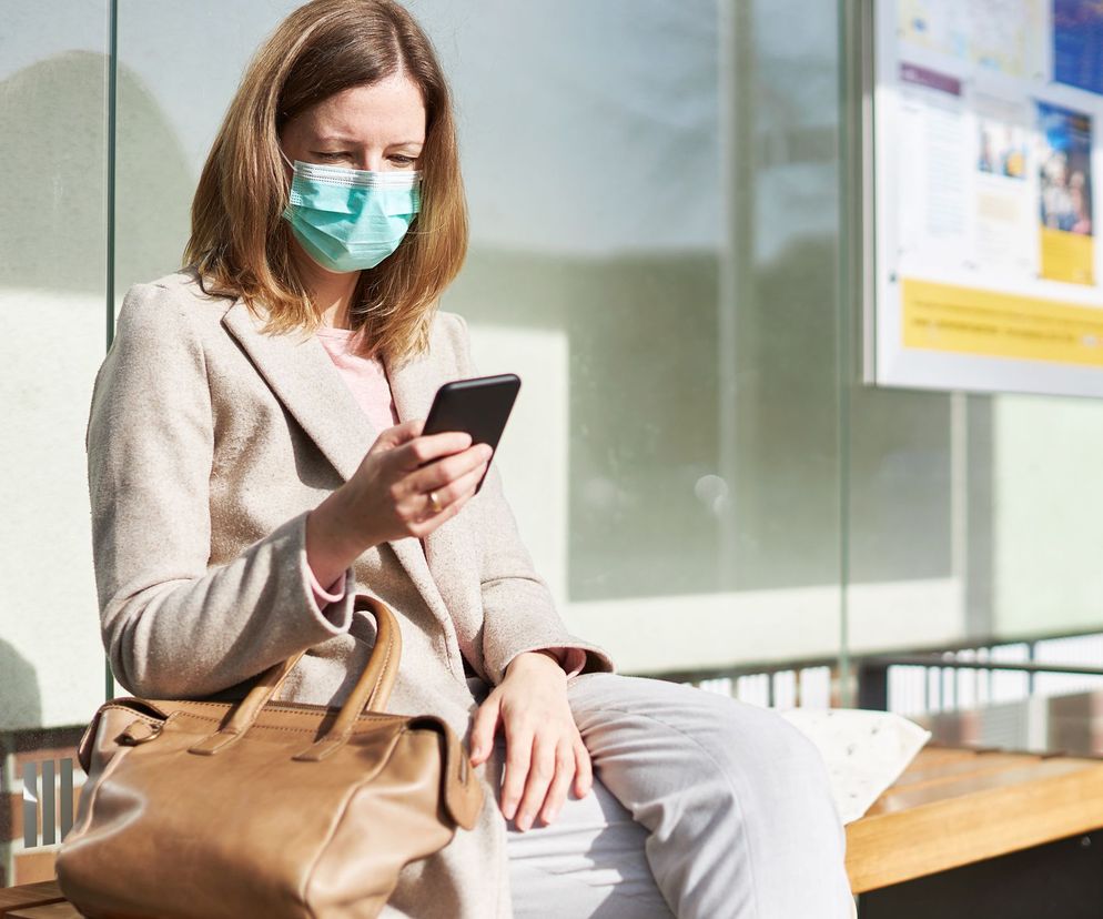 Kobieta w beżowym płaszczu i maseczce ochronnej na twarzy, siedząca na drewnianej ławce i wpatrująca się w smartfon. Obok niej brązowa torebka. Zdjęcie ilustruje temat dbania o higienę telefonu w kontekście koronawirusa. Więcej informacji znajdziesz na Poradnik Zdrowie.