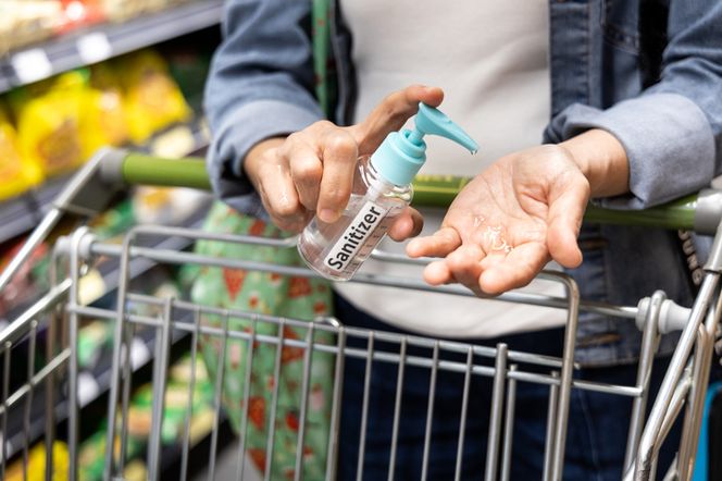 Osoba w dżinsowej kurtce dezynfekująca ręce żelem Sanitizer przy wózku sklepowym, symbolizująca dbałość o higienę i ostrzeżenia sanepidu, o których przeczytasz więcej na Poradnik Zdrowie.