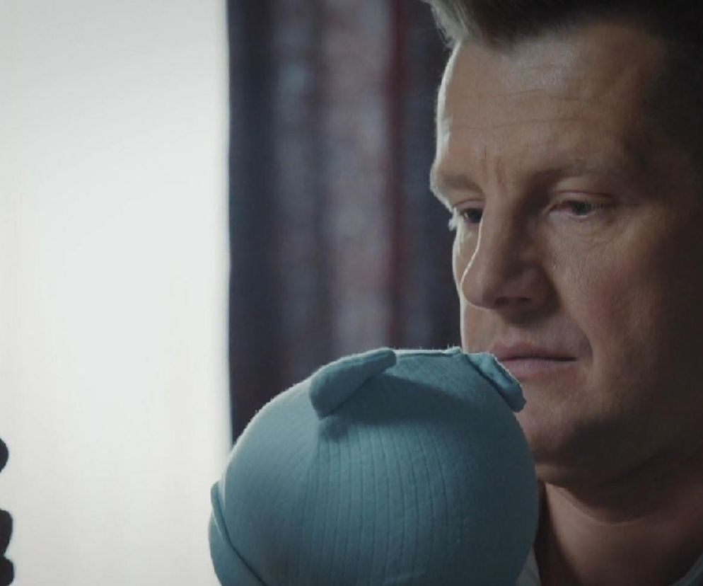 Paweł Zduński (Rafał Mroczek) z M jak miłość trzymający na rękach małego Antosia (Mark Mironienko) w błękitnej czapeczce. Scena oddaje emocje związane z opieką nad dzieckiem i relacje rodzinne, które możesz śledzić na Super Seriale.