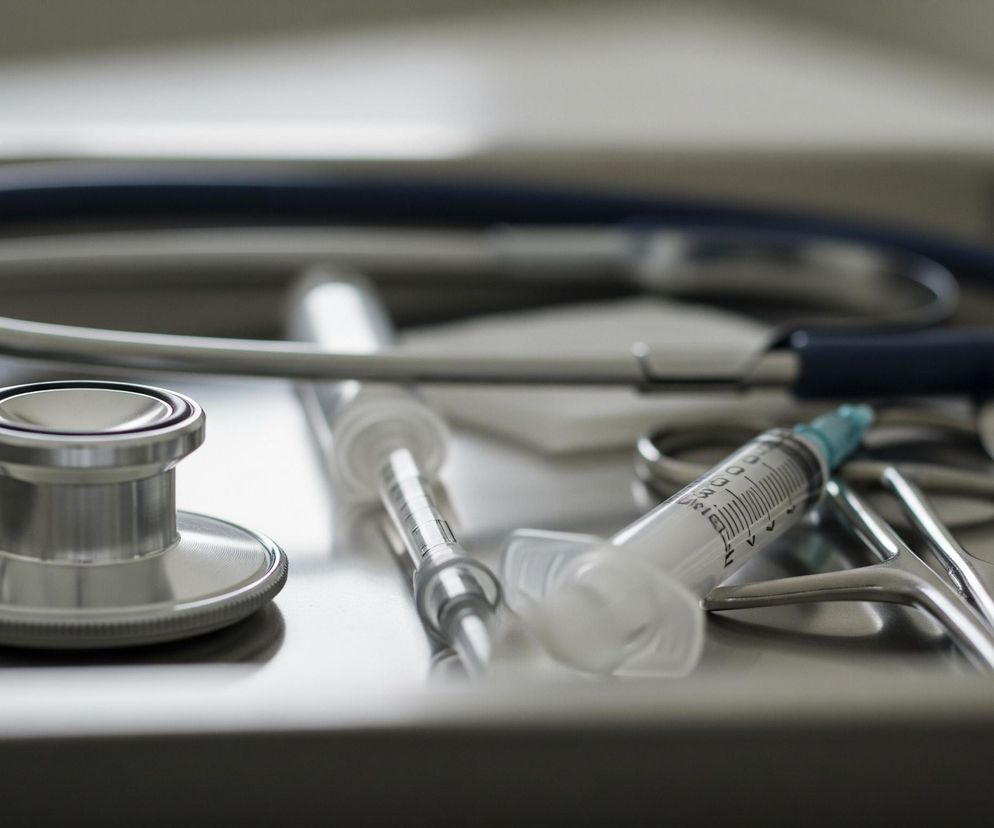 Stetoskop z błyszczącą metalową głowicą znajduje się na pierwszym planie po lewej stronie, z częścią ciemnoniebieskiego przewodu biegnącego w głąb kadru. Obok niego leży strzykawka z bezbarwnym korpusem i jasnoniebieską końcówką, a dalej widoczne są inne narzędzia chirurgiczne o metalicznym połysku, w tym nożyczki i prawdopodobnie kleszcze, ułożone na metalowej tacy. Tło jest rozmyte, utrzymane w jasnoszarych i białych tonach, z wyraźnym światłem odbijającym się od metalowych powierzchni.