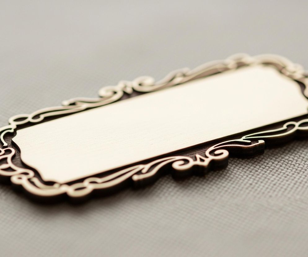 Prostokątna, beżowa tabliczka z zaokrąglonymi rogami, obramowana ciemnym, drewnianym wzorem z misternymi, ozdobnymi wycięciami przypominającymi wstążki lub arabeski. Tabliczka ma gładką, matową powierzchnię, a jej krawędzie są lekko podniesione, co tworzy trójwymiarowy efekt. Całość leży na jasnoszarym, teksturowanym tle, które ma widoczny splot tkaniny o drobnych, regularnych kwadratach. Oświetlenie pada z góry, tworząc subtelne cienie pod ozdobnym obramowaniem.