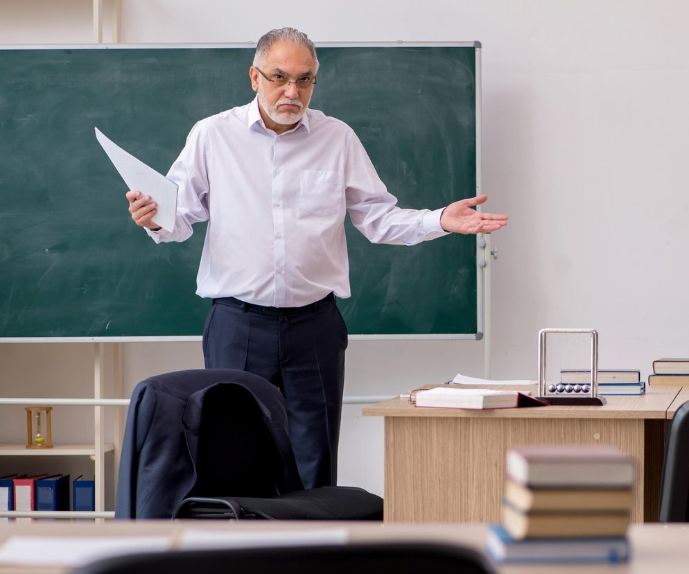 Starszy nauczyciel z brodą i w okularach, w białej koszuli, gestykuluje rękami, stojąc przed zieloną tablicą. W tle widoczny regał z kolorowymi segregatorami. Obraz symbolizuje wyzwania edukacji, o których możesz przeczytać na Super Biznes.