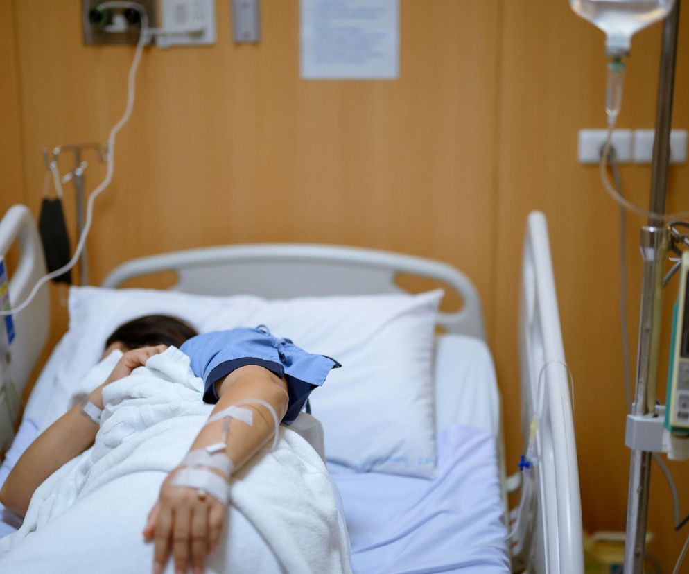 Pacjent leżący na szpitalnym łóżku podłączony do kroplówki, ubrany w niebieską koszulę, przykryty białym kocem. Fotografia oddaje trudną sytuację osób w szpitalach, nierzadko z powodu alkoholizmu, o czym przeczytasz więcej na Poradnik Zdrowie.