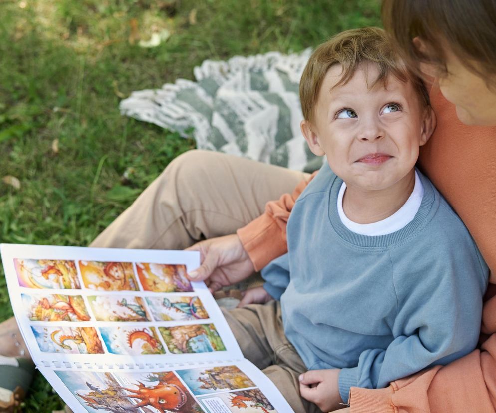 Uśmiechnięty chłopiec w niebieskiej bluzie z białą koszulką pod spodem, siedzący na kolanach matki, która czyta mu bajkę z ilustrowanej książki. Dziecko patrzy w górę, a matka pochyla się nad nim. Oboje siedzą na trawie. Więcej o dziedziczeniu inteligencji na Poradnik Zdrowie.