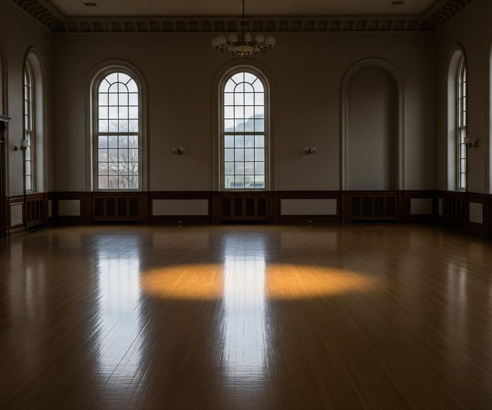 Przestronne, puste pomieszczenie z lśniącą, drewnianą podłogą w ciepłym odcieniu brązu. Na podłodze widoczne są intensywne, prostokątne refleksy światła, z czego środkowy ma wyraźnie żółto-pomarańczową barwę, a pozostałe są białe i rozproszone. Ściany są kremowo-beżowe, z ciemniejszymi, drewnianymi boazeriami w dolnej części, a w górnej części ozdobione są rzędami półokrągłych okien z kratami, przez które widać niewyraźny zimowy krajobraz z drzewami i budynkami. Sufit zdobi rzeźbiony gzyms, a pośrodku pomieszczenia wisi żyrandol z wieloma ramionami.