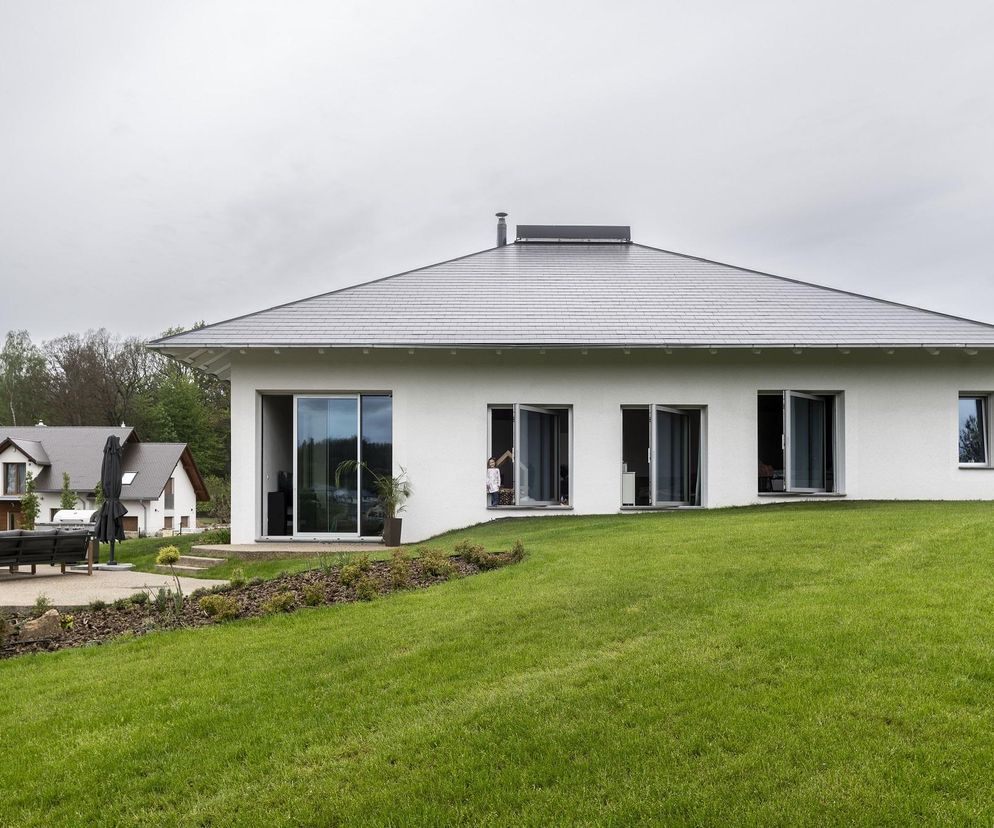 Biały jednorodzinny Dom w Staniszowie, z szarym spadzistym dachem i dużymi przeszkleniami wychodzącymi na zielony, pochyły trawnik. Architektura Murator Plus prezentuje projekt XM Architekten, Karol Łącki i Dominik Pierzchlewicz, zgłoszony do konkursu Życie w Architekturze 2025.
