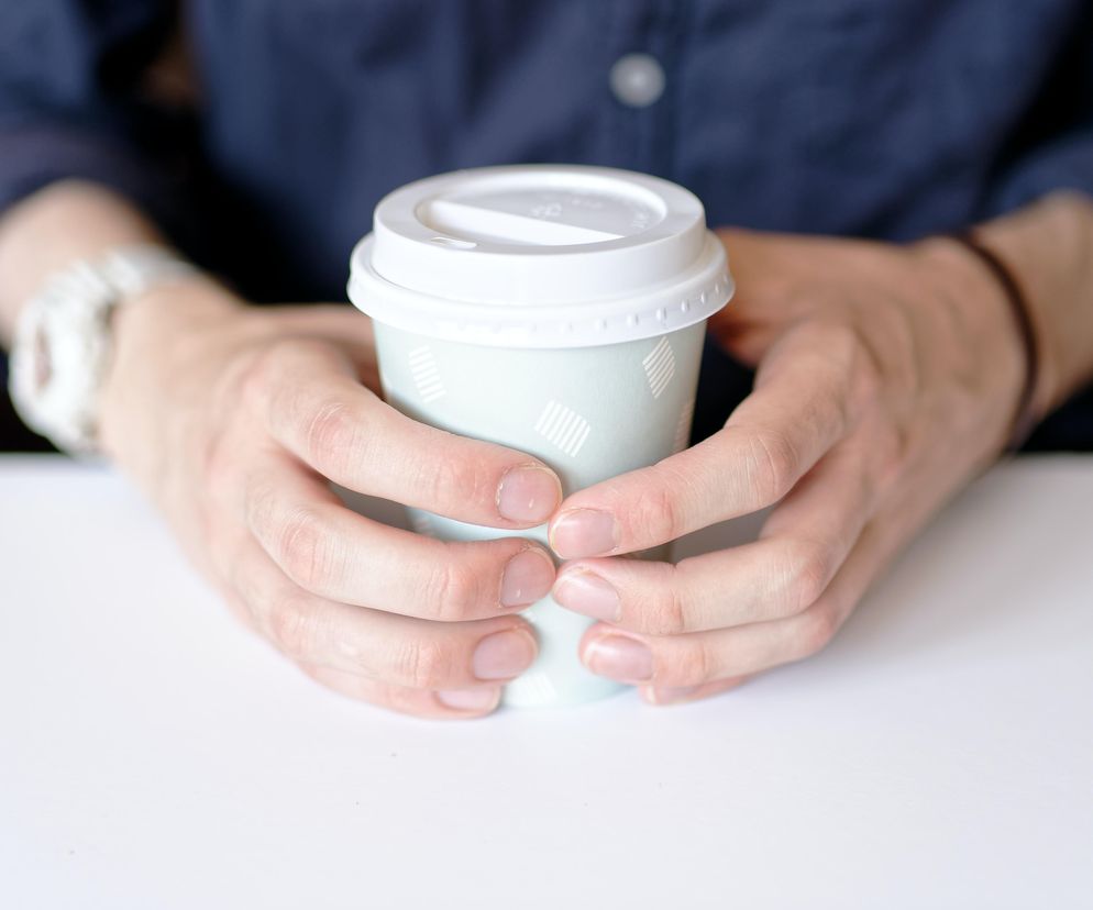 Dłonie osoby trzymającej papierowy kubek na kawę z białą przykrywką, symbolizujące problem z recyklingiem opakowań wielomateriałowych. O tym, gdzie należy wyrzucić takie kubki, przeczytasz na Super Biznes.