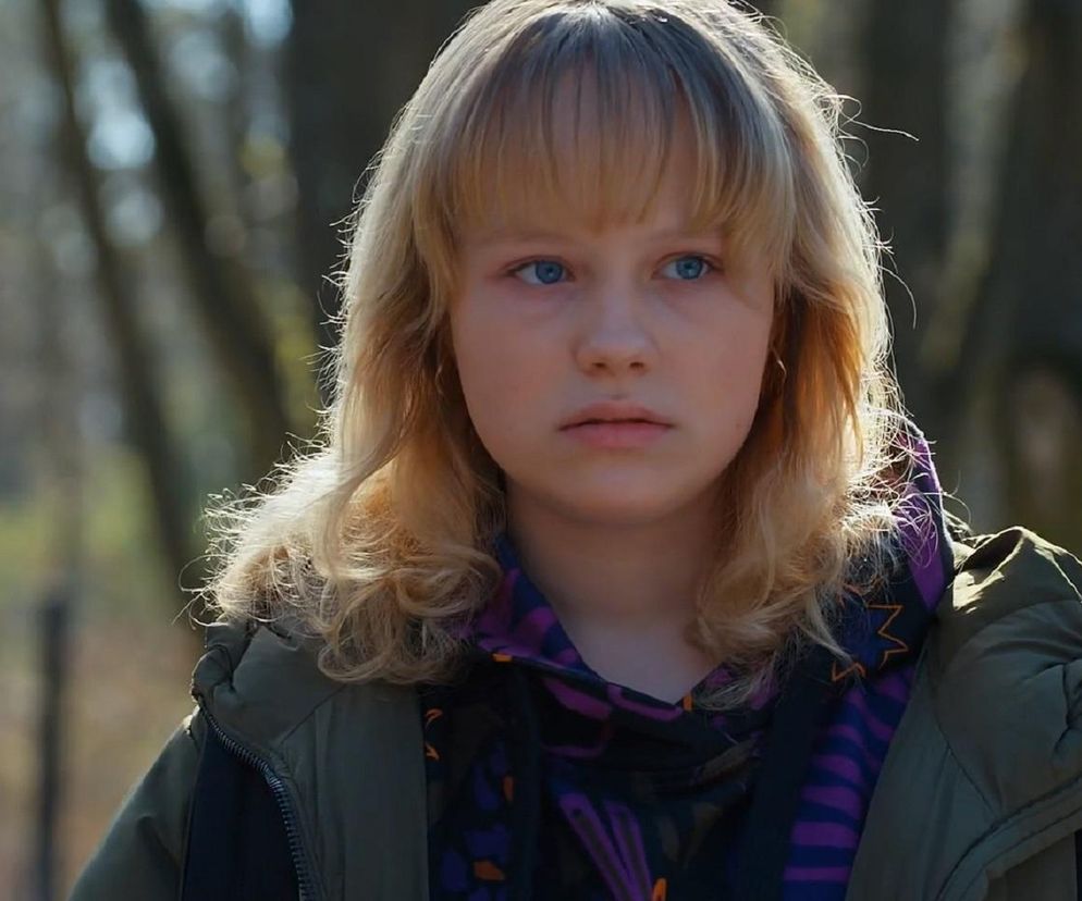 Młoda, blondwłosa Emilka (Jessika Frankiewicz) z niebieskimi oczami, w kurtce khaki i fioletowej bluzie z wzorami, wygląda na zmartwioną. Tło stanowi rozmyty las, co podkreśla jej samotność. Więcej o losach Emilki przeczytasz na Super Seriale.