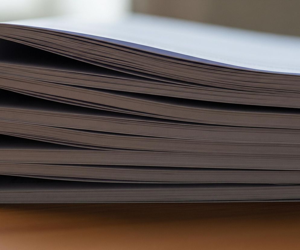 Na pierwszym planie widoczny jest stos czterech zamkniętych, nieoprawionych notatników lub zeszytów ułożonych jeden na drugim, leżących na drewnianej powierzchni. Ich krawędzie są delikatnie zaokrąglone, a wewnętrzne strony stron są ciemniejsze, co tworzy wyraźny kontrast z jaśniejszymi zewnętrznymi krawędziami. Strona prawa góra stosu jest nieznacznie wygięta do góry, co sugeruje, że leżą one płasko na powierzchni. Całość kadru jest oświetlona z góry, tworząc subtelne cienie i akcentując teksturę papieru.