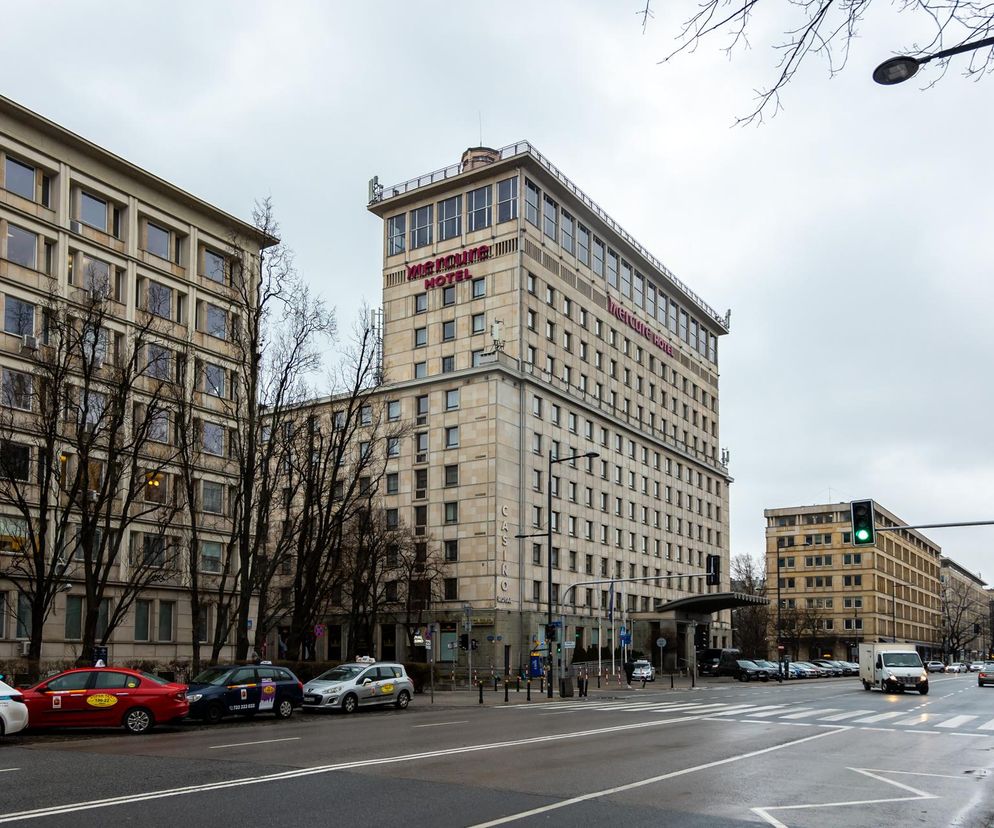 Hotel Mercure Warszawa Grand przy ulicy Kruczej, widziany z ulicy, z czerwonymi napisami MERCURE HOTEL na fasadzie. Budynek o jasnej elewacji, z charakterystycznym daszkiem nad wejściem, po prawej stronie oświetlona loggia kawiarni. Obok taksówki i drzewa bez liści, pod pochmurnym niebem. Więcej o historii Grand Hotelu w Warszawie przeczytasz na Architektura Murator Plus.