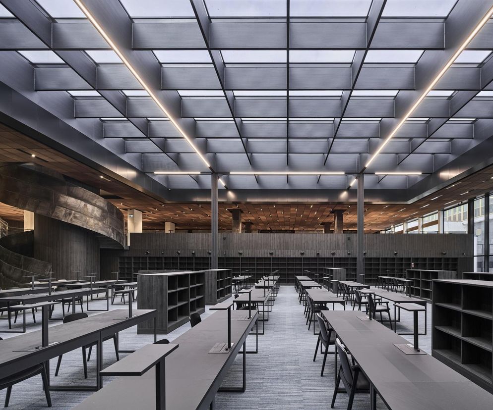 Wnętrze Biblioteki Narodowej z nowoczesną, przestronną czytelnią dolną. Widoczne długie stoły, wygodne krzesła, regały na książki, spiralna klatka schodowa z mosiądzu oraz świetlik dachowy zapewniający naturalne światło. Architektura Murator Plus to portal, gdzie można dowiedzieć się więcej o tym projekcie.