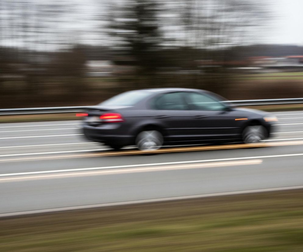 Ciemny samochód osobowy pędzi po wielopasmowej drodze w lewą stronę kadru, z rozmytym tłem sugerującym dużą prędkość. Auto jest w kolorze granatowym lub ciemnoszarym, z widocznymi czerwonymi światłami tylnymi oraz pomarańczowym światłem kierunkowskazu z przodu. Na drodze, wyznaczone są białe pasy ruchu oraz dwa żółte pasy oddzielające jezdnie, również rozmyte. Na lewej krawędzi drogi znajduje się srebrna barierka energochłonna, za którą widać rozmyte zielono-brązowe pobocze, a dalej ciemne, rozmyte drzewa i zabudowania wiejskie z czerwonymi dachami.