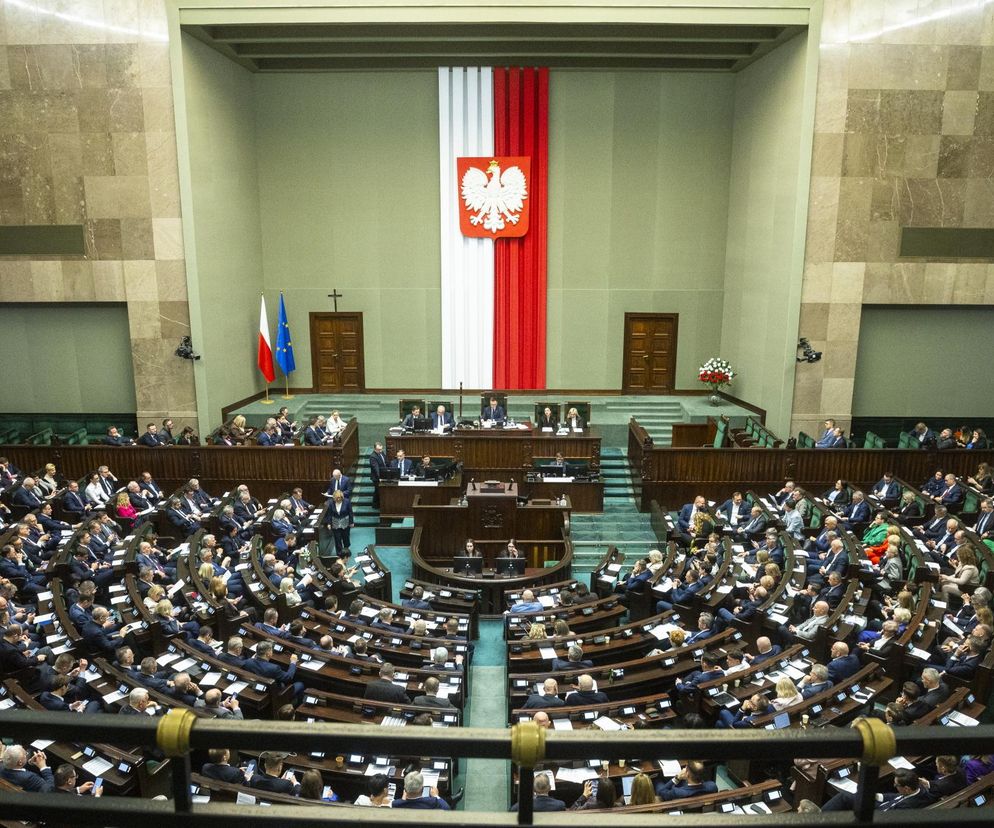 Wypełnioną posłami salę plenarną Sejmu, gdzie parlamentarzyści obradują nad kluczowymi ustawami dotyczącymi Polaków, w tym bonem ciepłowniczym i Kodeksem pracy. W tle widoczny polski orzeł na biało-czerwonej fladze oraz flaga Unii Europejskiej. Przebieg tego intensywnego posiedzenia Sejmu śledź na Super Biznes, by być na bieżąco z polityczną bombą w parlamencie.