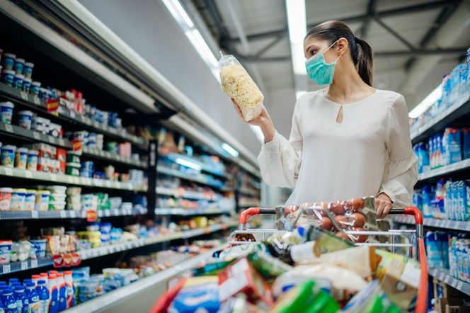 Kobieta w maseczce ochronnej podczas zakupów w supermarkecie, trzymająca w ręku opakowanie produktu. Ilustracja symbolizuje zagrożenie zdrowia, o którym można przeczytać na portalu Poradnik Zdrowie.