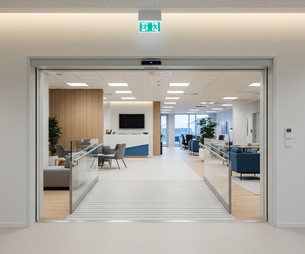 Przez otwarte, automatyczne drzwi, składające się z dwóch ruchomych paneli ze szkła i metalu, widać nowoczesne wnętrze biurowe. W centralnej części obrazu znajduje się jasna posadzka z poziomymi, ciemnymi liniami, prowadząca do dalszej części pomieszczenia z beżową i drewnianą podłogą, oraz ciemnoniebieskimi fotelami. Nad drzwiami widoczny jest zielony znak wyjścia ewakuacyjnego, a po prawej stronie, na białej ścianie, znajduje się podświetlony symbol osoby w garniturze w okręgu. W głębi, po lewej, za szklaną barierką z metalową poręczą, znajdują się szare fotele i drewniane panele ścienne.