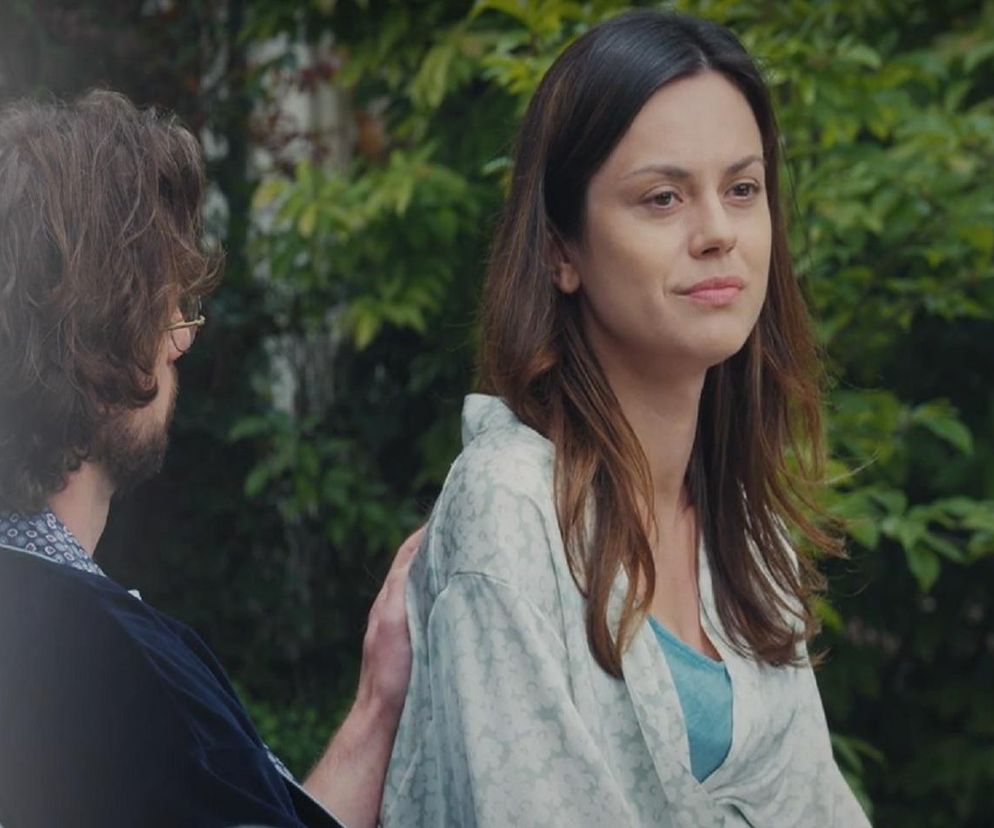 Paulina Lasota jako Kasia, ubrana w jasną narzutkę, z długimi brązowymi włosami, siedzi obok Jakuba Karskiego (Krzysztof Kwiatkowski), który kładzie jej dłoń na ramieniu w tle zielonych liści. Więcej o ich losach w M jak miłość na portalu Super Seriale.