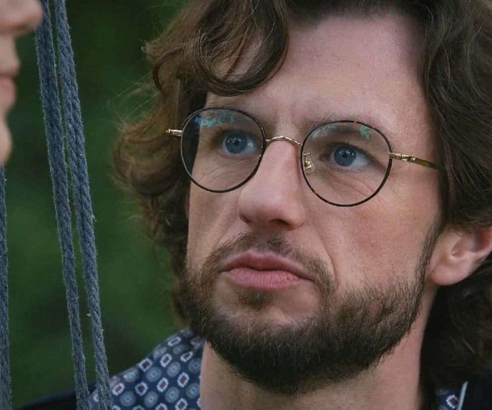 Mariusz (Mateusz Mosiewicz) w okrągłych okularach, z bujnymi włosami i brodą, patrzy w górę, wyrażając zaskoczenie lub oburzenie. Fragment twarzy Kasi (Paulina Lasota) z warkoczykami widoczny z lewej. Scena konfliktu w serialu M jak miłość na portalu Super Seriale.