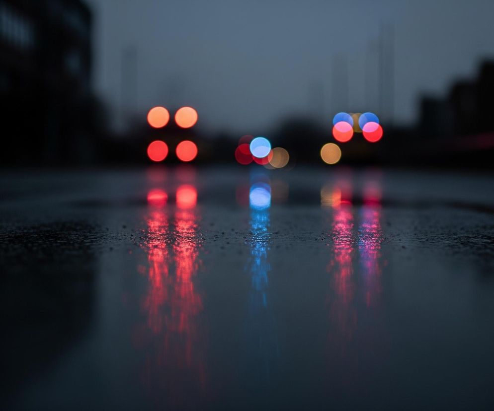 Na mokrej, asfaltowej nawierzchni odbijają się rozmyte światła samochodów. Dominują tu czerwień i pomarańcz tylnych świateł oraz sporadyczne, jasnoniebieskie światła przednie. Tło jest bardzo niewyraźne, zarysowują się na nim ciemne, zamazane kształty budynków lub innych konstrukcji pod szarym, zachmurzonym niebem. Całość kompozycji koncentruje się na refleksach świetlnych na drodze, tworząc atmosferę miejskiej nocy lub zmierzchu.