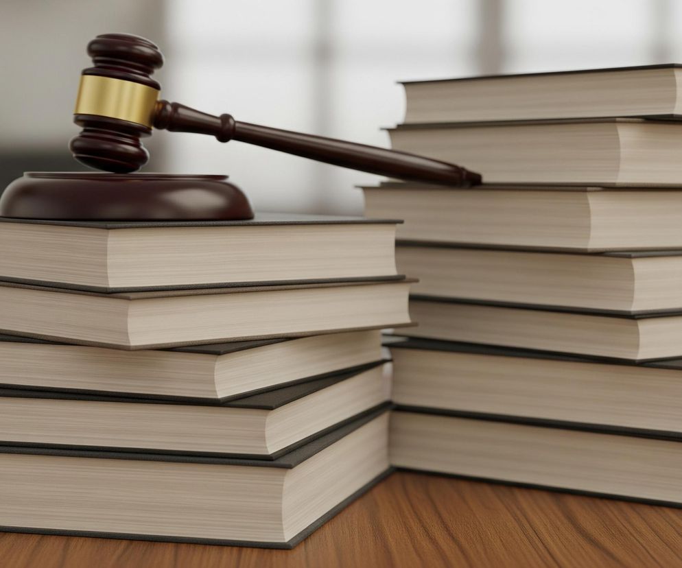 Na drewnianym blacie stołu leży brązowy młotek sędziowski ze złotą obręczą, ułożony na ciemnej podstawce. Obok młotka znajdują się dwie ułożone na sobie stosy książek w ciemnych oprawach z jasnymi kartkami. Lewy stos składa się z czterech, a prawy z pięciu książek. Tło jest rozmyte, utrzymane w jasnych, neutralnych barwach.