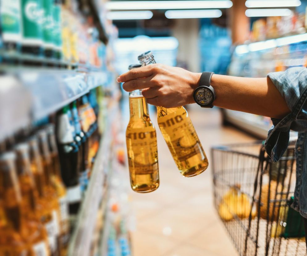 Ręka trzymająca dwie butelki piwa w supermarkecie symbolizująca dostępność alkoholu. Zdjęcie ilustruje temat zakazu sprzedaży alkoholu na stacjach paliw i w uzdrowiskach, o czym szerzej można przeczytać na portalu Poradnik Zdrowie.