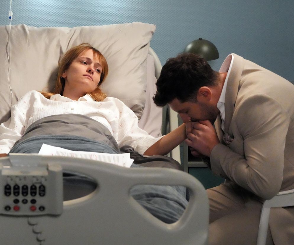 Michalina Robakiewicz jako Ola, leży w szpitalnym łóżku w białej koszuli i szarym kocu, z aparaturą medyczną przy poduszce. Obok niej, Jasper Sołtysiewicz jako Justin, pochyla się, całując jej dłoń. Zdjęcie ilustruje dramatyczne wydarzenia z serialu Barwy Szczęścia na portalu Super Seriale.