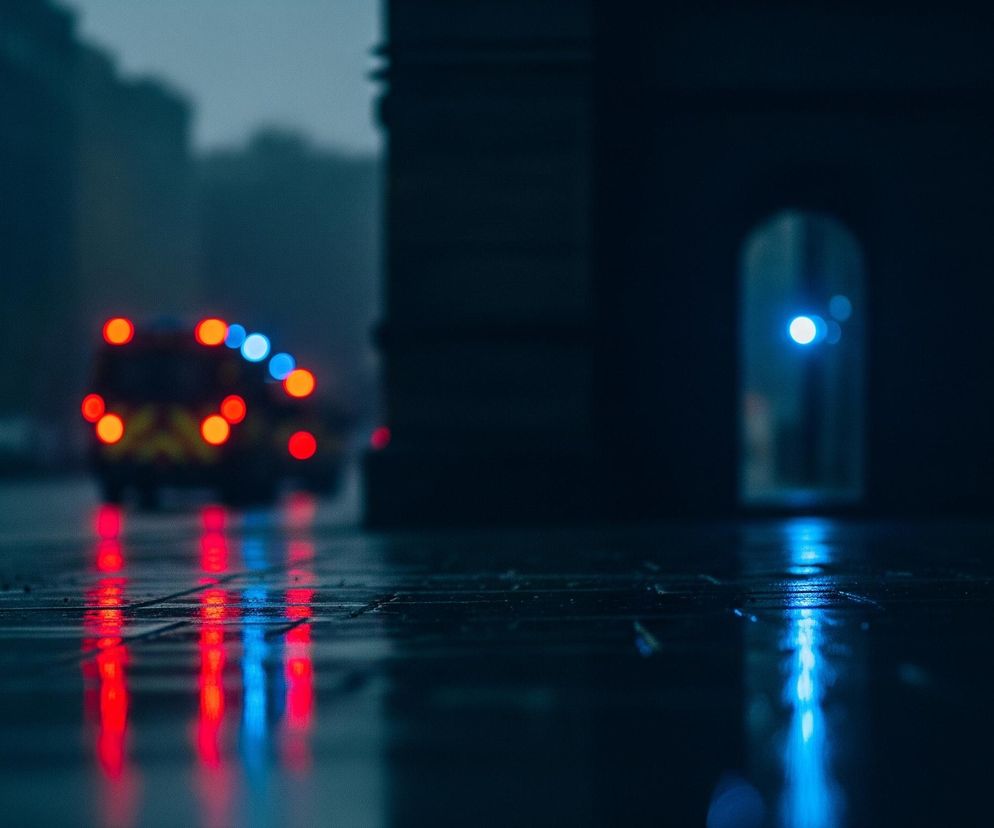 Uliczny krajobraz o zmierzchu, z mokrą nawierzchnią drogi odbijającą światła. W centrum, na drodze widoczny jest samochód służb ratunkowych, którego tylne czerwone światła i niebieskie sygnalizacyjne są jaskrawo rozświetlone. Po prawej stronie, w oddali, widać budynek z łukowatym przejściem, w którym również świeci się niebieskie światło. Całość utrzymana jest w ciemnych, chłodnych barwach, dominują odcienie granatu i szarości.