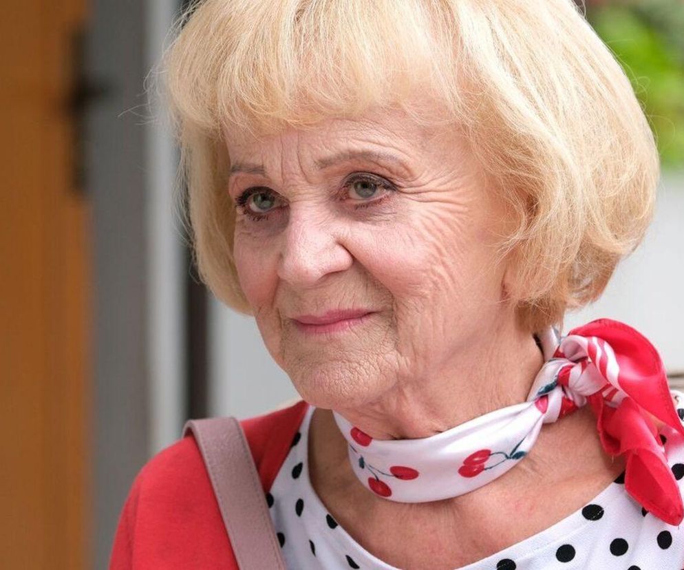 Zofia Kisielowa z serialu M jak miłość z krótkimi blond włosami i delikatnym makijażem, uśmiechnięta, w czerwonym kardiganie i białej bluzce w grochy, z apaszką w wisienki na szyi. W tle niewyraźny budynek. O zmianach w jej życiu przeczytasz na Super Seriale.