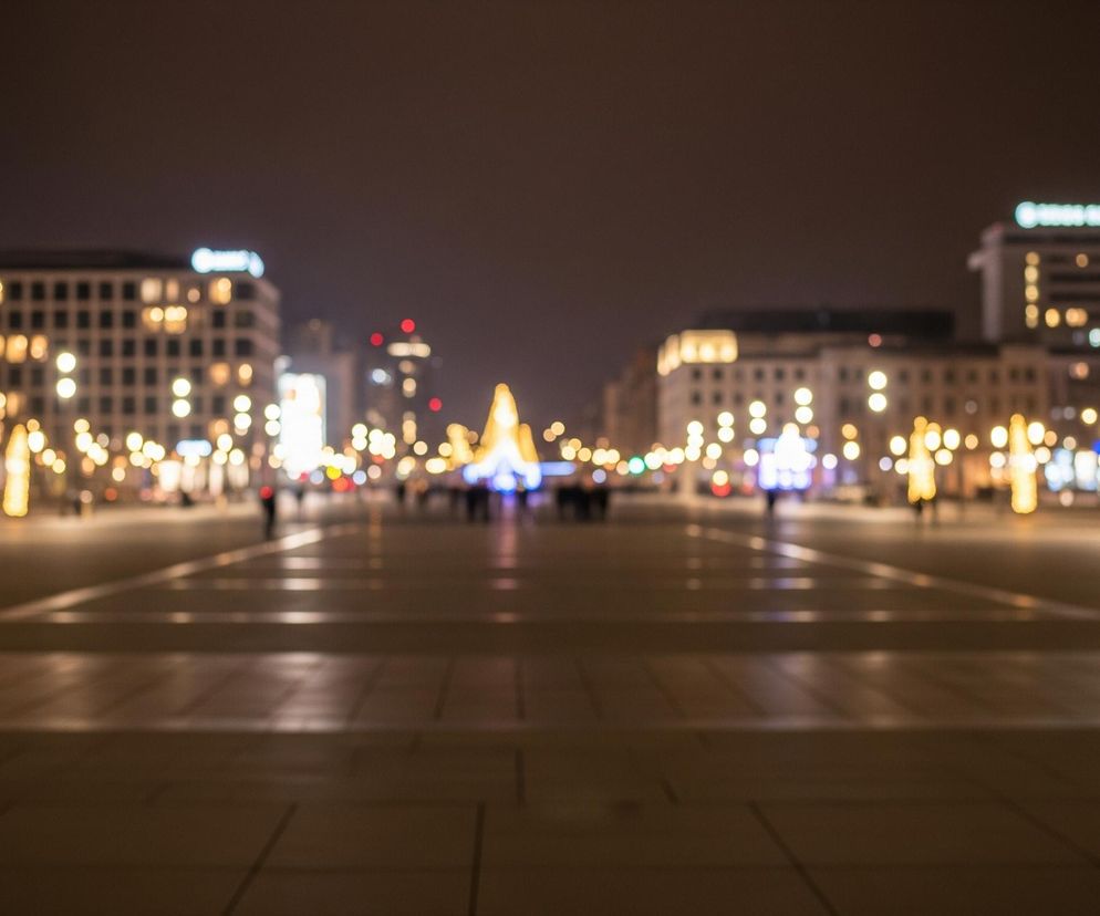 Duży plac miejski nocą, oświetlony licznymi, rozmazanymi światłami w ciepłych, żółto-pomarańczowych barwach. W tle widoczne są kontury budynków z podświetlonymi oknami, a na horyzoncie majaczy rozświetlona, stożkowata struktura, prawdopodobnie choinka. Na placu można dostrzec niewyraźne sylwetki ludzi oraz jasne refleksy na mokrej nawierzchni, tworzące poziome pasy światła. Całość utrzymana jest w ciemnych odcieniach brązu i czerni, z dominującymi akcentami jasnych punktów świetlnych, nadającymi scenerii świąteczny lub wieczorny charakter.