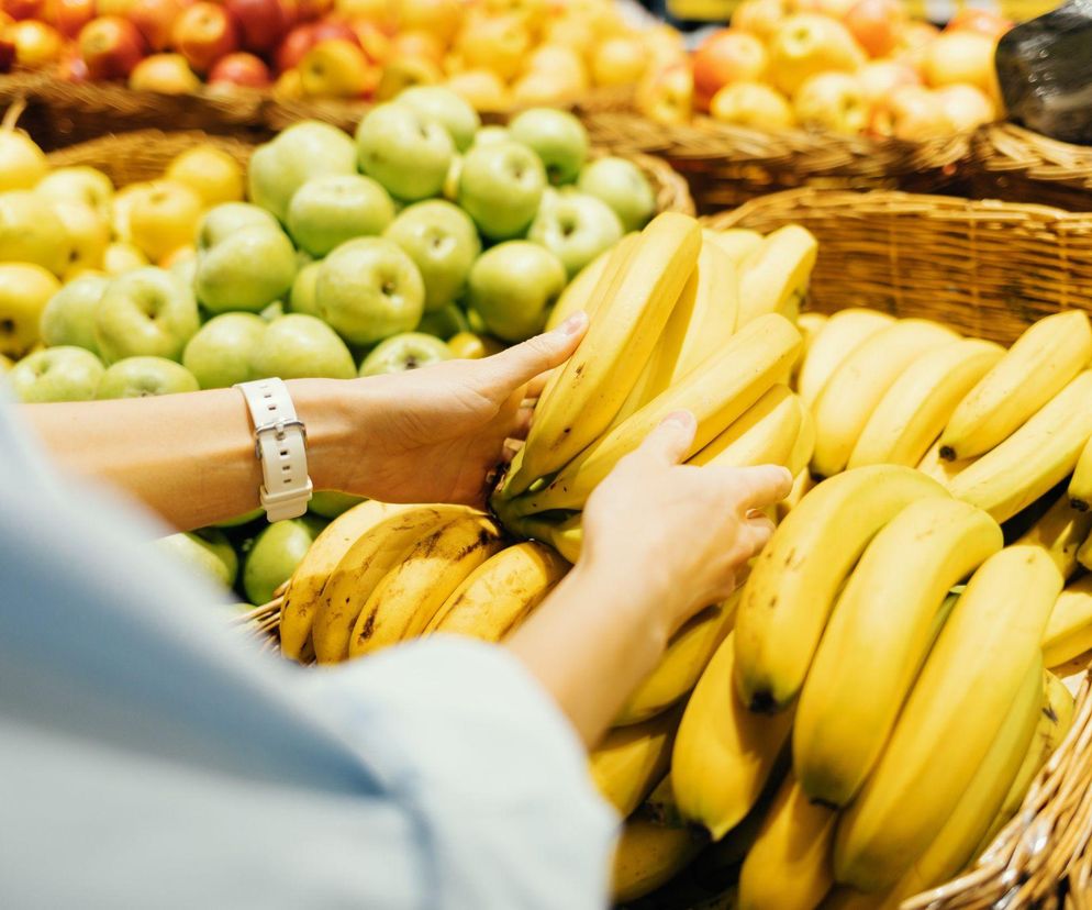 Kobieta w jasnej bluzce i białym zegarku wybiera świeże banany z wiklinowego kosza w supermarkecie. W tle widać kosze pełne zielonych i czerwonych jabłek. Dowiedz się więcej o zdrowej diecie na Poradnik Zdrowie.