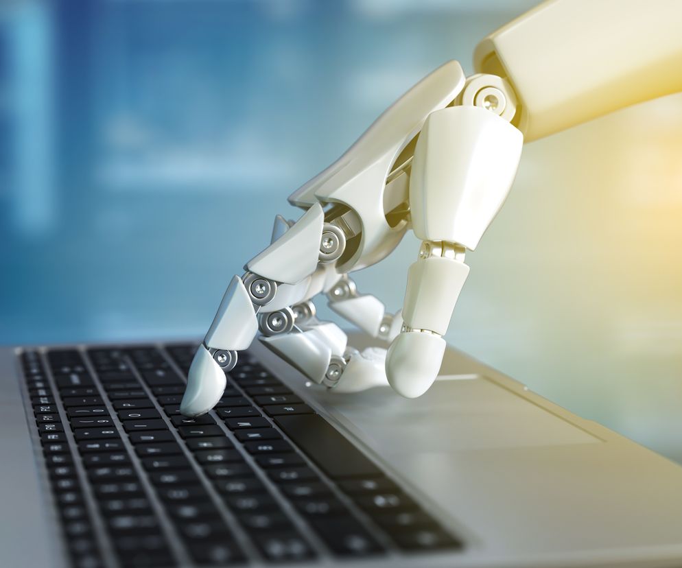 Mechaniczna dłoń robota z jasnego metalu naciska klawisz na klawiaturze laptopa, symbolizując automatyzację procesów i przyszłość technologii w medycynie. Artykuł na Poradnik Zdrowie opisuje zastosowanie robotów w szpitalach.