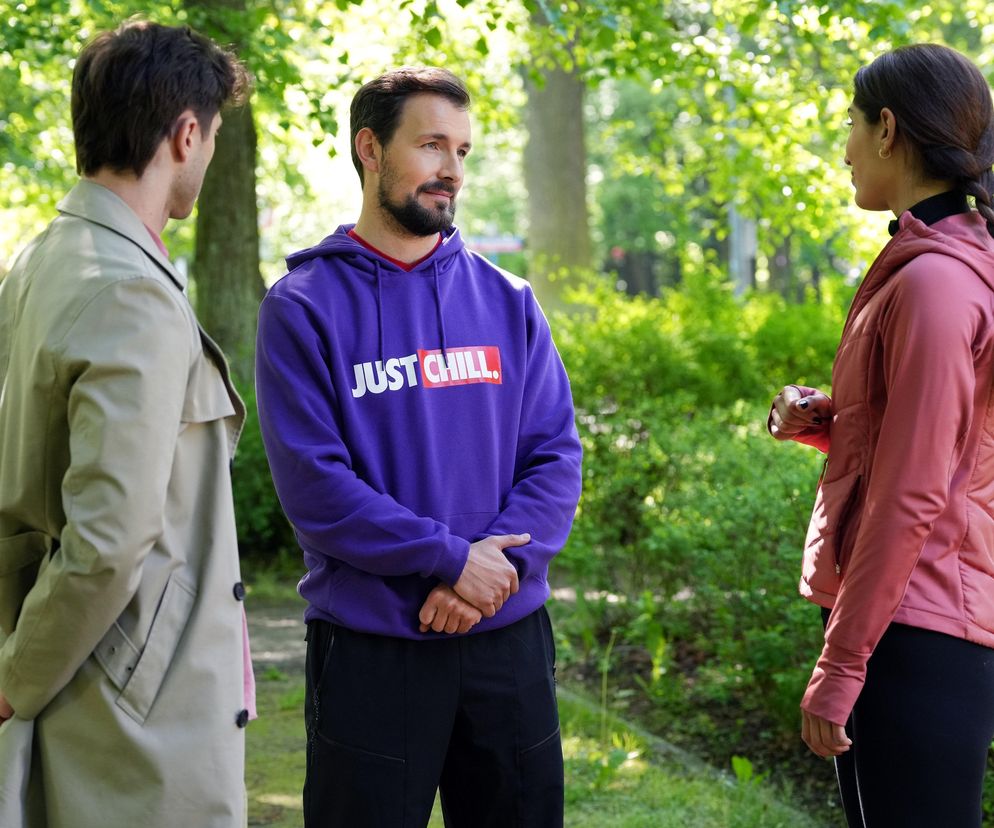 Trzy osoby, dwie mężczyzny i kobieta, rozmawiają w parku. Jeden z mężczyzn w fioletowej bluzie z napisem JUST CHILL. i kobieta z warkoczem w różowej kurtce patrzą na trzeciego mężczyznę w beżowym płaszczu. Można przeczytać o tym więcej na Super Seriale.