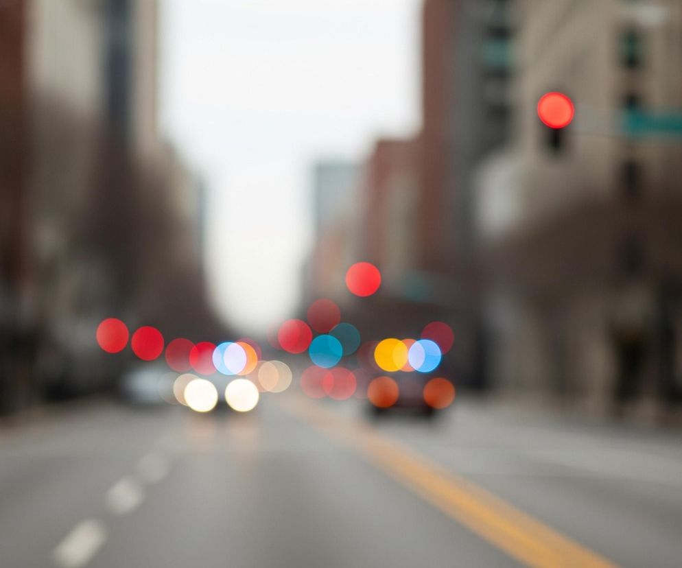 Rozmyty widok miejskiej ulicy, skupiając się na efekcie bokeh świateł. W centrum kadru widać szarą nawierzchnię drogi z rozmytą żółtą linią ciągłą po prawej stronie i przerywanymi białymi liniami po lewej. Nad drogą unoszą się liczne, kolorowe kręgi świetlne: dominują czerwone, symbolizujące prawdopodobnie światła stopu lub sygnalizację, a także jasne, białe i żółte, reprezentujące przednie światła samochodów oraz niebieskie i turkusowe. Po lewej stronie, na pierwszym planie, widoczne jest jedno wyraźne, czerwone światło, prawdopodobnie sygnalizator. W tle zarysy budynków rozciągają się wzdłuż obu stron ulicy, ich detale są jednak całkowicie zatarte.