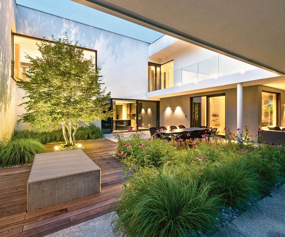 Nowoczesne patio z drewnianym tarasem, zielonymi trawami ozdobnymi i kwitnącymi różowymi kwiatami. Podświetlone drzewko i część wypoczynkowa z hamakiem. Na naszym portalu znajdziesz więcej inspiracji.
