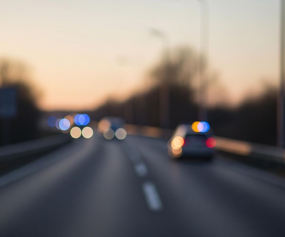 Rozmyta scena przedstawia drogę o zmierzchu lub świcie. Centralnie biegnie dwupasmowa droga, z widocznymi przerywanymi białymi liniami. Po prawej stronie drogi, w oddali, widać zarys samochodu z niebieskim i pomarańczowym światłem na dachu oraz czerwonymi światłami z tyłu. Z przodu, na drugim pasie, widoczne są rozmyte, jasne światła nadjeżdżających pojazdów, głównie białe i niebieskie. Horyzont zdominowany jest przez rozmyte kształty drzew i krzewów, a nad nimi rozciąga się niebo o barwach przechodzących od jasnego beżu do delikatnego pomarańczu.
